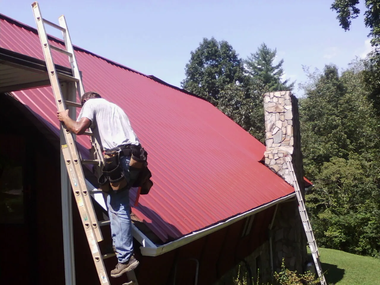 Roofer on a ladder beside a red metal roof and stone chimney — Soffit & Fascia Repair by Roofing Brothers in Minburn