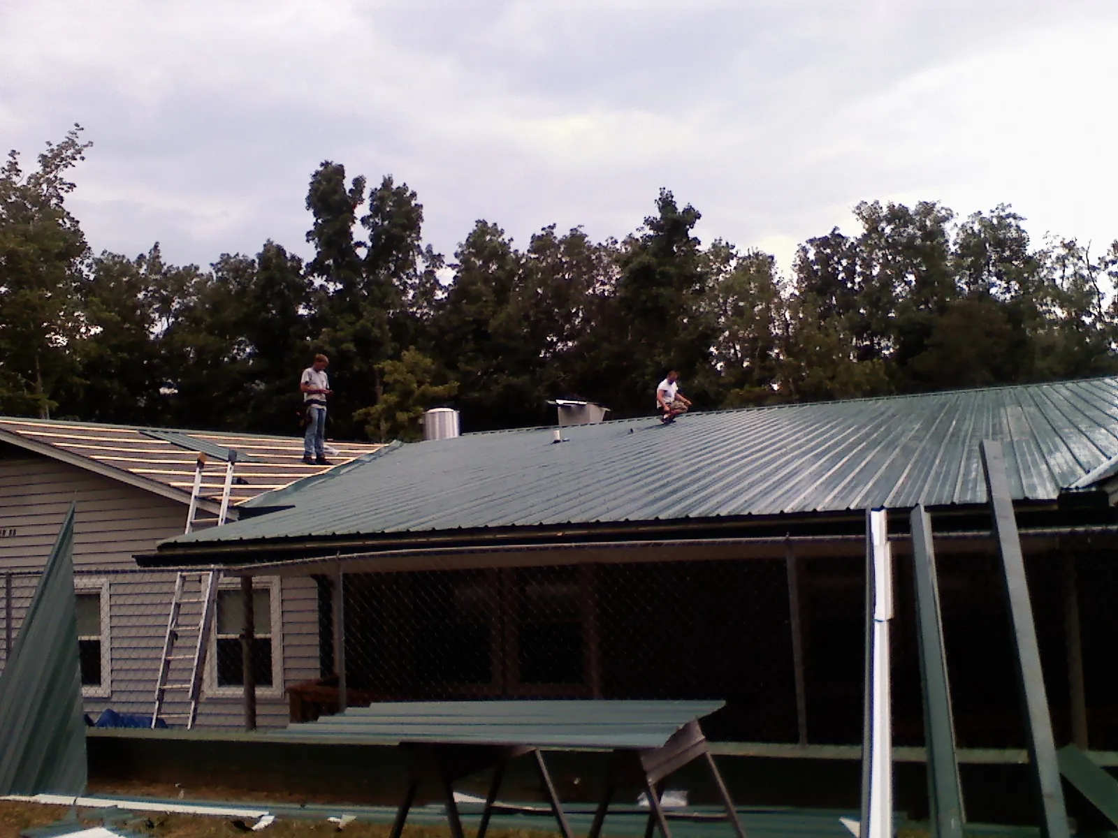 Two roofers installing green metal panels above a covered porch in Minburn